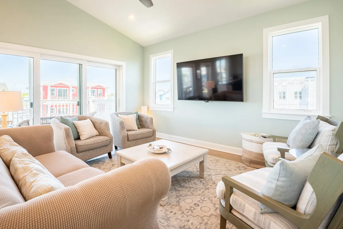 A comfortable seating area with plush sofas faces a large wall-mounted television, all bathed in natural light from expansive windows. The arrangement creates an inviting space for group relaxation, conversation, or watching movies together.