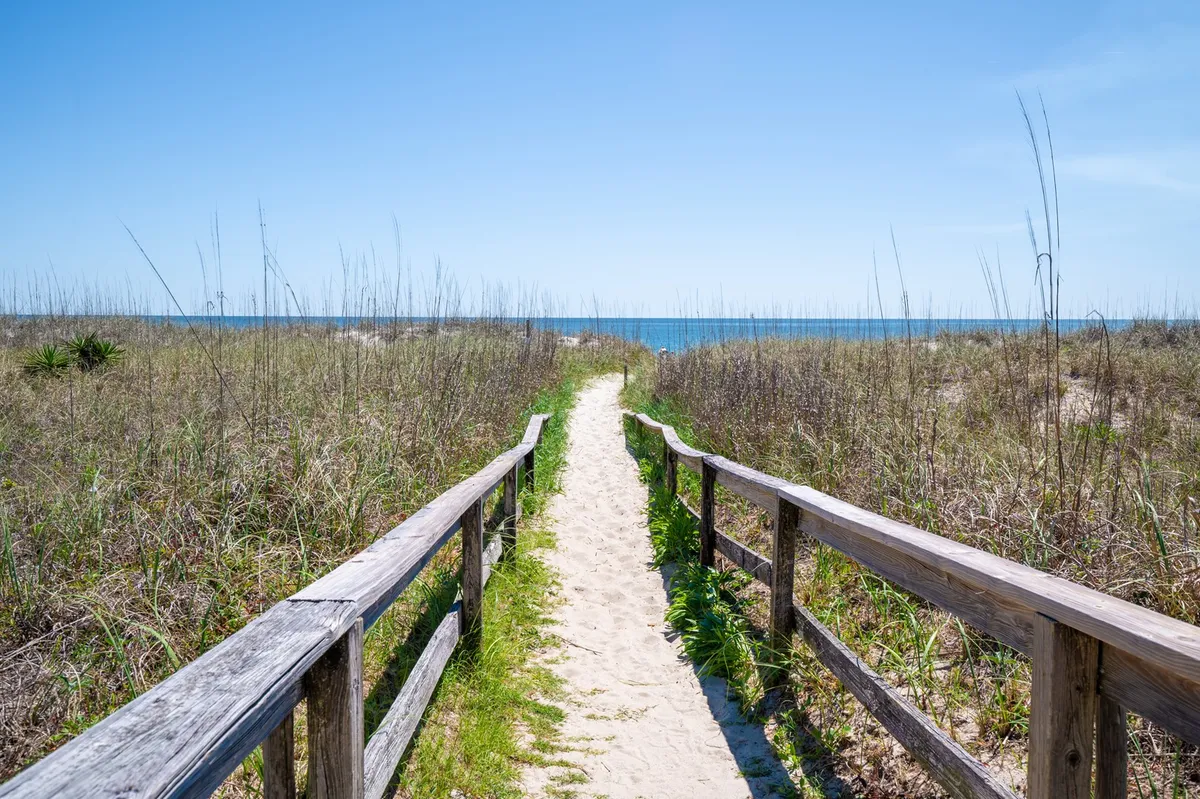 A wooden boardwalk winds through dune grass, leading directly to a sandy beach with a sweeping view of the ocean and sky. The scene is tranquil, with the soft textures of sand and vegetation underscoring the proximity to the water.