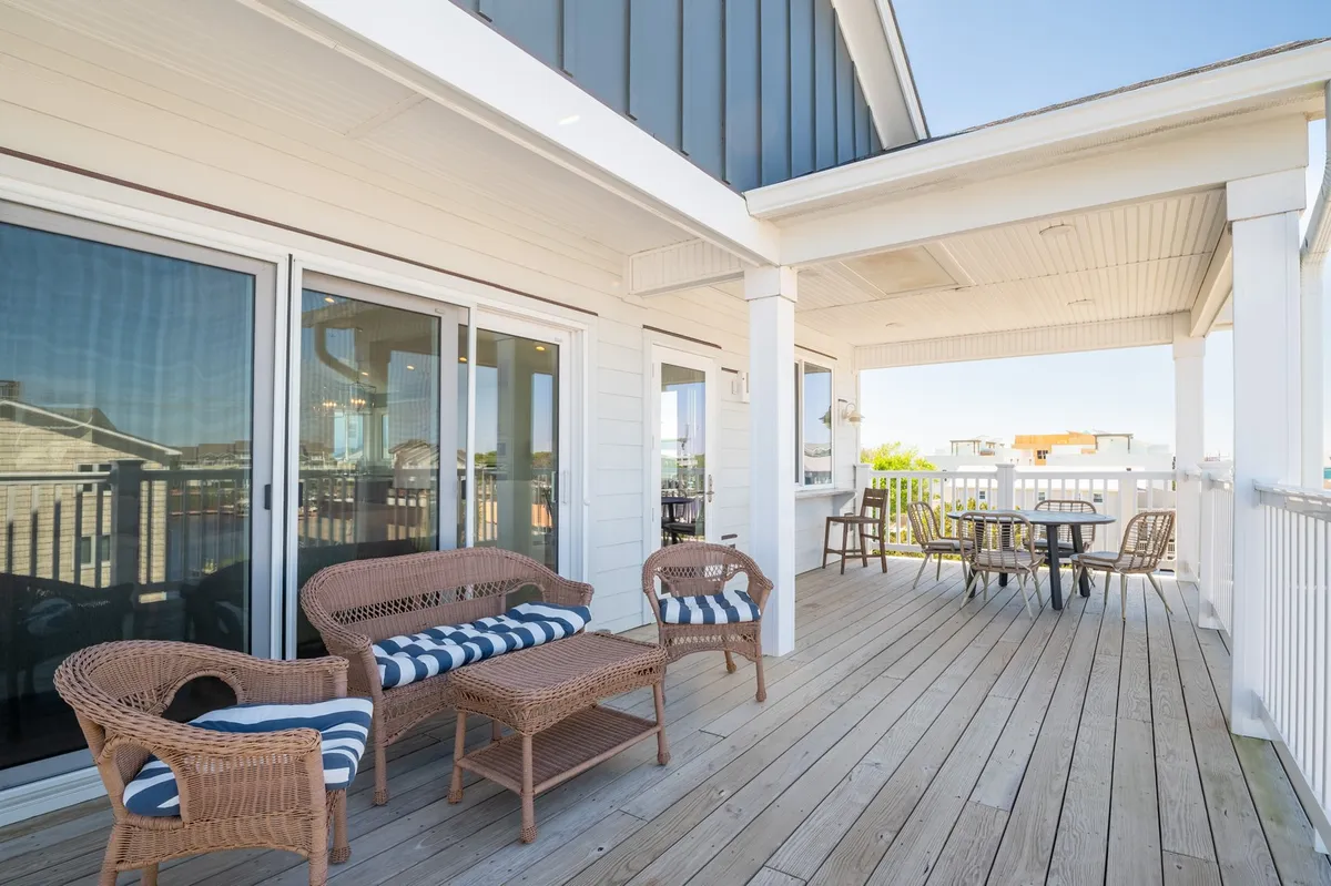 This outdoor deck scene showcases wicker chairs and a wooden table positioned near a sturdy railing, with large glass doors leading back into the house. The setup is perfect for relaxing or entertaining outdoors while enjoying water or sunset views.