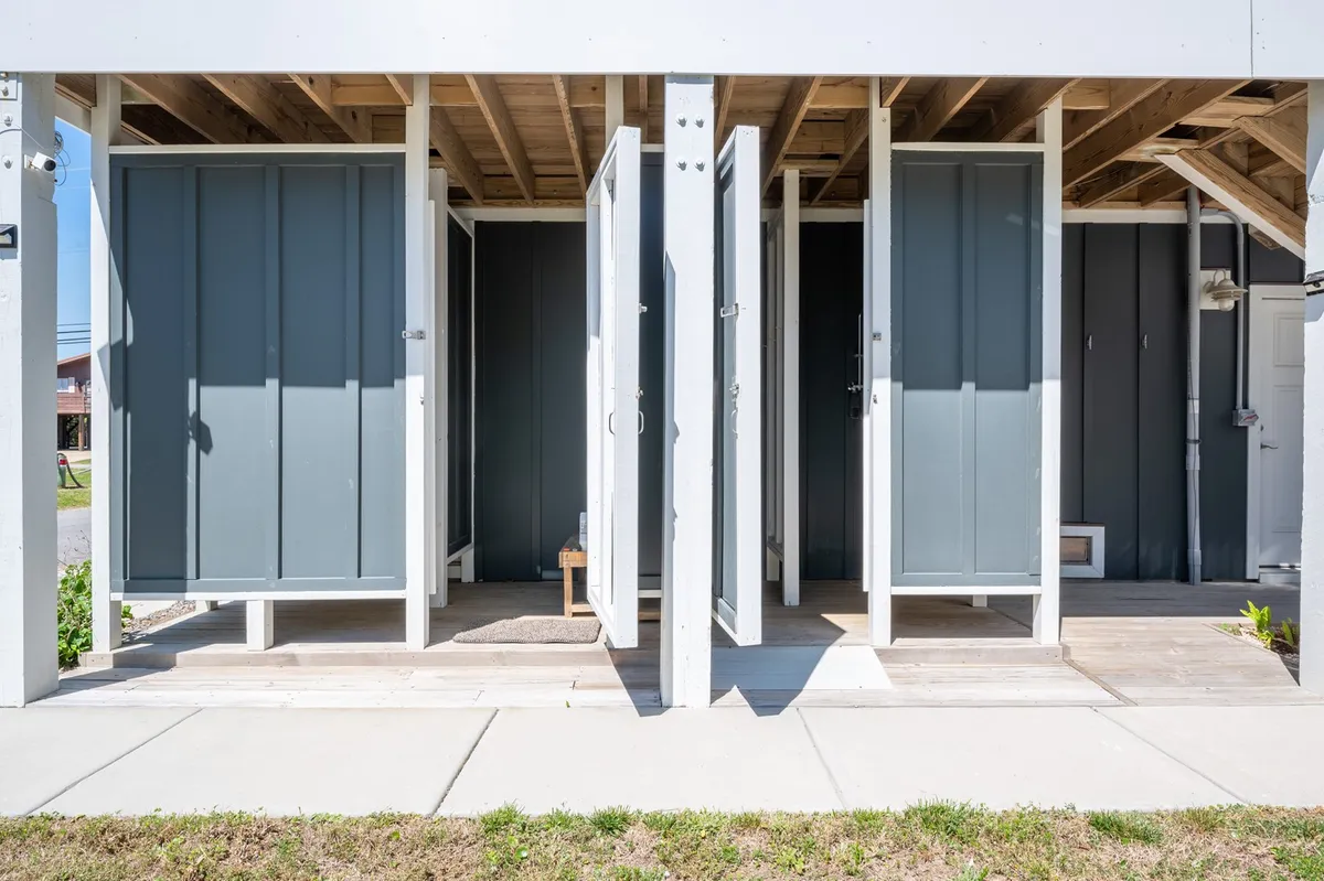 A bright exterior shot of dual outdoor shower stalls set on a wooden deck, with a visible pathway leading from the showers toward the yard. The setup suggests a convenient spot to rinse off after beach trips, supporting active, sandy afternoons.