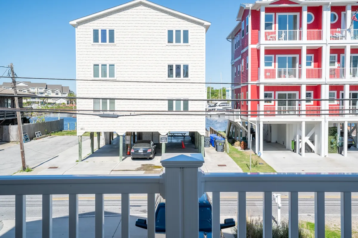 A 'view' image from a balcony, showing two neighboring buildings, a parking area, and a glimpse of the water beyond. The perspective places viewers physically and thematically between home and the wider world, hinting at easy access to exploration and nearby attractions.