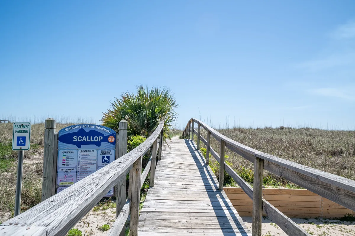 A wooden boardwalk with a welcoming sign is surrounded by lush greenery, leading toward the shore. The entrance to the beach is inviting and accessible, hinting at the easy journey from the home to the ocean.