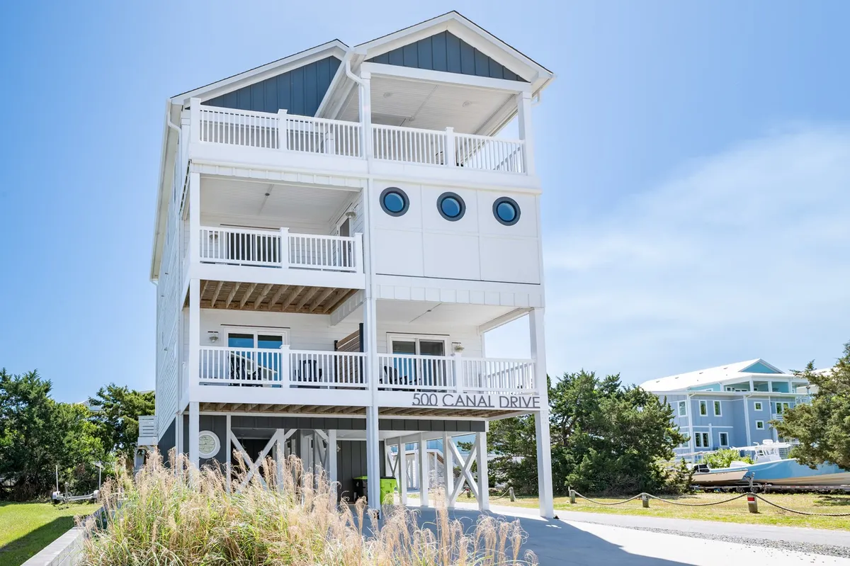 The image showcases the exterior of Dietrich Beach House at Carolina Beach, North Carolina. The home features signature three circular windows, an expansive deck area perfect for outdoor dining and relaxation, and a prominent sign with the property's address. The deck captures beautiful sunsets and offers a space to sip morning coffee while enjoying ocean and marina views, all just steps from the beach.