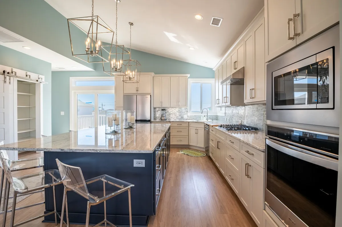 This image features a spacious, modern kitchen with a prominent granite island at the center, surrounded by hanging light fixtures and stainless steel appliances. Large windows frame beautiful natural light, hinting at water views beyond. The open layout encourages cooking, entertaining, and mingling.