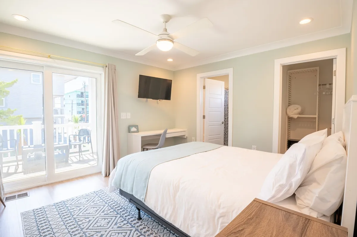 A calm and spacious bedroom with a large bed, balcony door, desk, and television. The room is filled with soft natural light, offering a relaxing and versatile retreat.