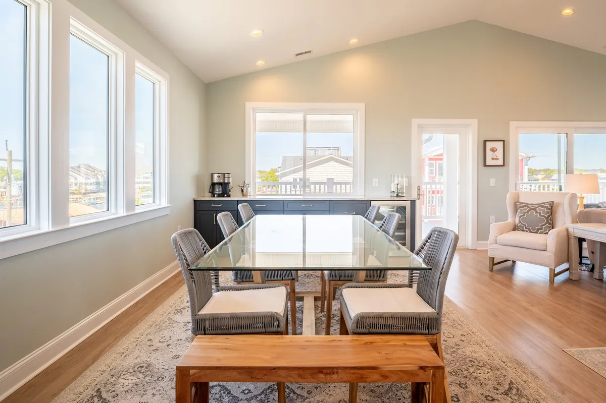 This image displays a bright, sun-filled dining area with a glass-top table surrounded by comfortable seating, set beside large windows that bathe the space in natural light. The ambiance is warm and welcoming, ideal for family meals or game nights.