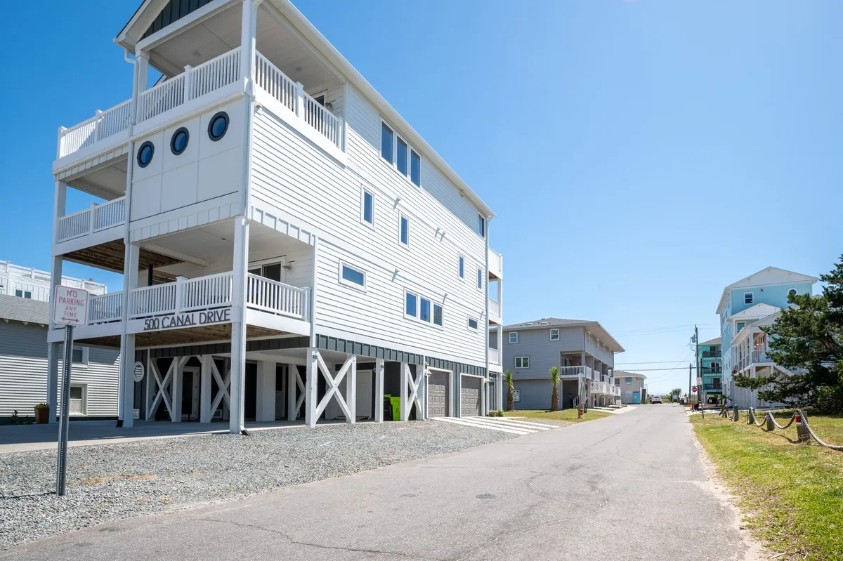 A modern beach house exterior featuring a welcoming front porch with seating, large windows, and a coastal architectural style. The front porch hints at a friendly, walkable neighborhood and a relaxing base for adventures.