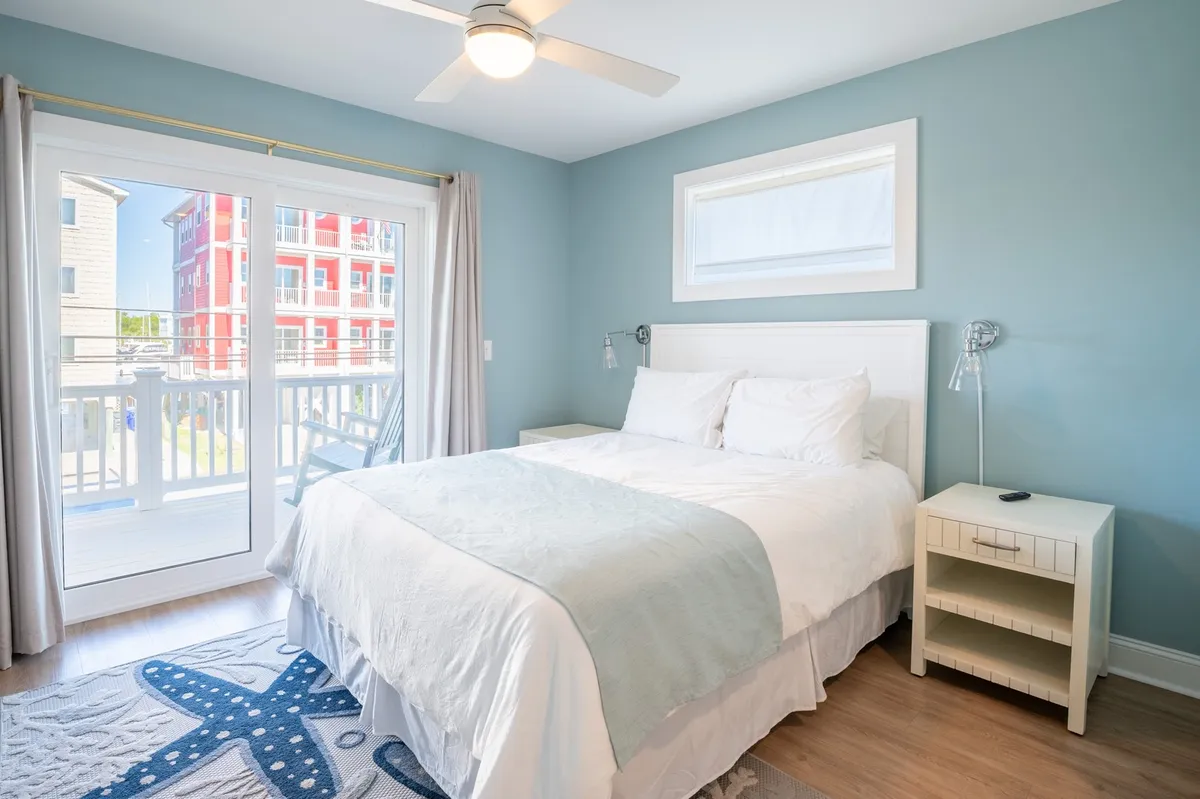 A serene bedroom featuring a plush bed with white linens and blue accents, complemented by a beach-themed area rug. Large sliding glass doors lead to a private balcony, bathing the room in natural light.