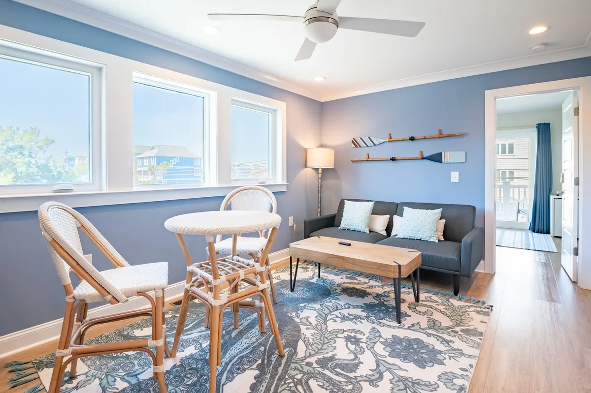 A cozy lounge area featuring a bistro table and chairs, a plush sofa with decorative pillows, large windows letting in ample natural light, and a decorative wall shelf. The space exudes warmth and versatility, suitable for casual gatherings or quiet relaxation.